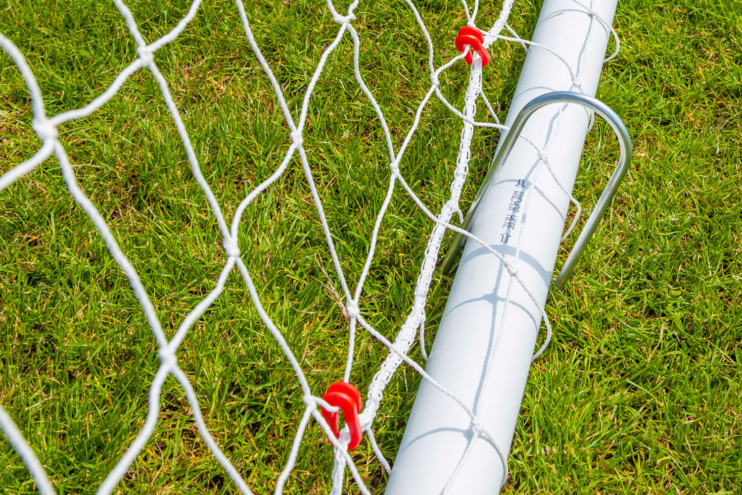 Close-up of the Samba Sports Samba Match Goal 8 x 6, showing its durable white uPVC frame on green grass, red clips securing the netting, and a metal stake fixing the goal firmly to the ground.