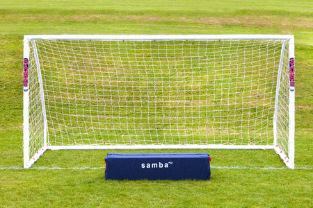 A Samba Sports Match Goal 12 x 6 stands on a grassy field with a net. In front is a dark blue rectangular bag labeled "Samba Match Goal" in white text. The weather is clear and bright.