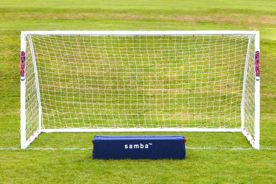 A Samba Sports Match Goal 12 x 6 stands on a grassy field with a net. In front is a dark blue rectangular bag labeled "Samba Match Goal" in white text. The weather is clear and bright.