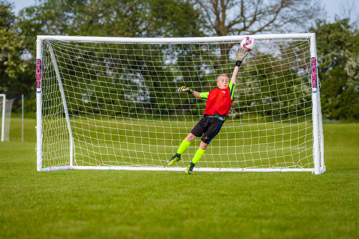 A soccer goalkeeper in a red jersey and yellow socks leaps right, fully outstretched, trying to block a ball headed for the top corner of a Samba Sports Samba Match Goal 8 x 6 on a grassy field with trees in the background.