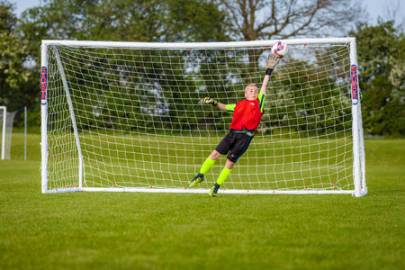 A soccer goalkeeper in a red jersey and yellow socks leaps right, fully outstretched, trying to block a ball headed for the top corner of a Samba Sports Samba Match Goal 8 x 6 on a grassy field with trees in the background.