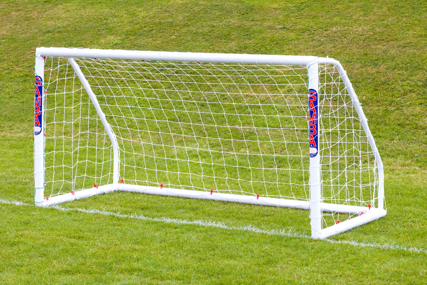 The Samba Sports Samba Match Goal 8 x 4, an easy-to-assemble white football goal with "SAMBA" printed vertically on its posts, stands with its net on a grassy field against a gently sloped background.