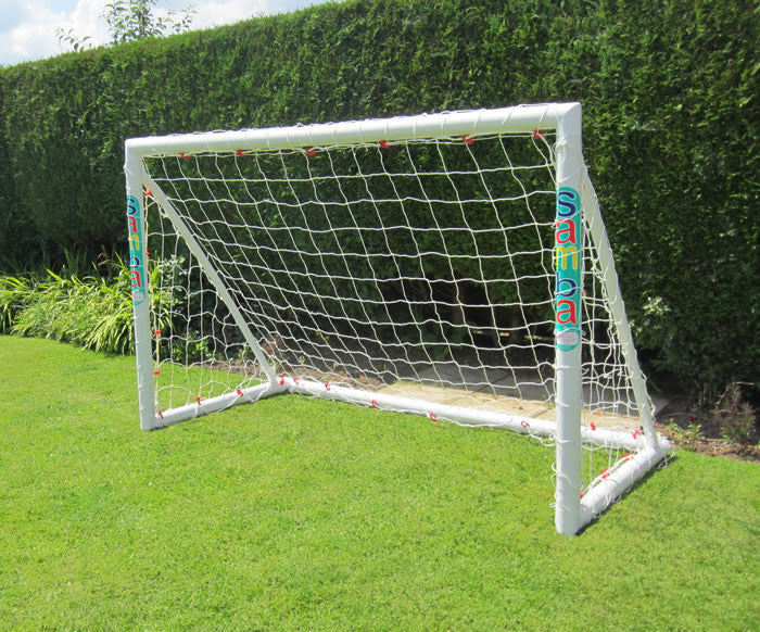 A Samba Sports Trainer Goal 6 x 4, a portable white uPVC football goal with net, stands on a green lawn. "Samba" is printed on its sides, with tall hedges and plants behind it under a partly cloudy sky.