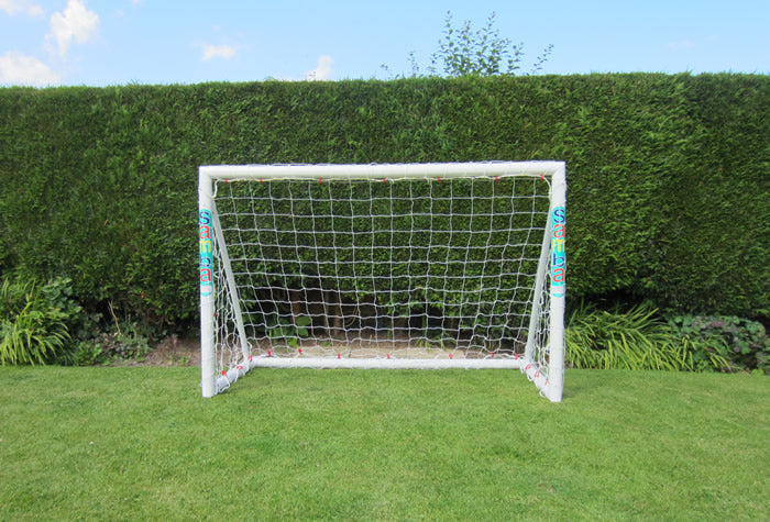 A Samba Sports Trainer Goal 6 x 4, made from white uPVC with a net, stands on green grass in front of a tall, trimmed hedge under a blue sky.