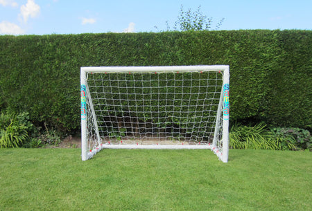 A Samba Sports Trainer Goal 6 x 4, made from white uPVC with a net, stands on green grass in front of a tall, trimmed hedge under a blue sky.
