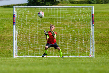 A young soccer goalie in a red and green uniform stands ready in front of a Samba Sports Samba Futsal Match Goal 3m x 2m as a soccer ball approaches on a grassy field.