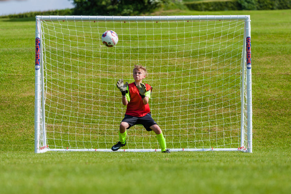 A young soccer goalie in a red and green uniform stands ready in front of a Samba Sports Samba Futsal Match Goal 3m x 2m as a soccer ball approaches on a grassy field.