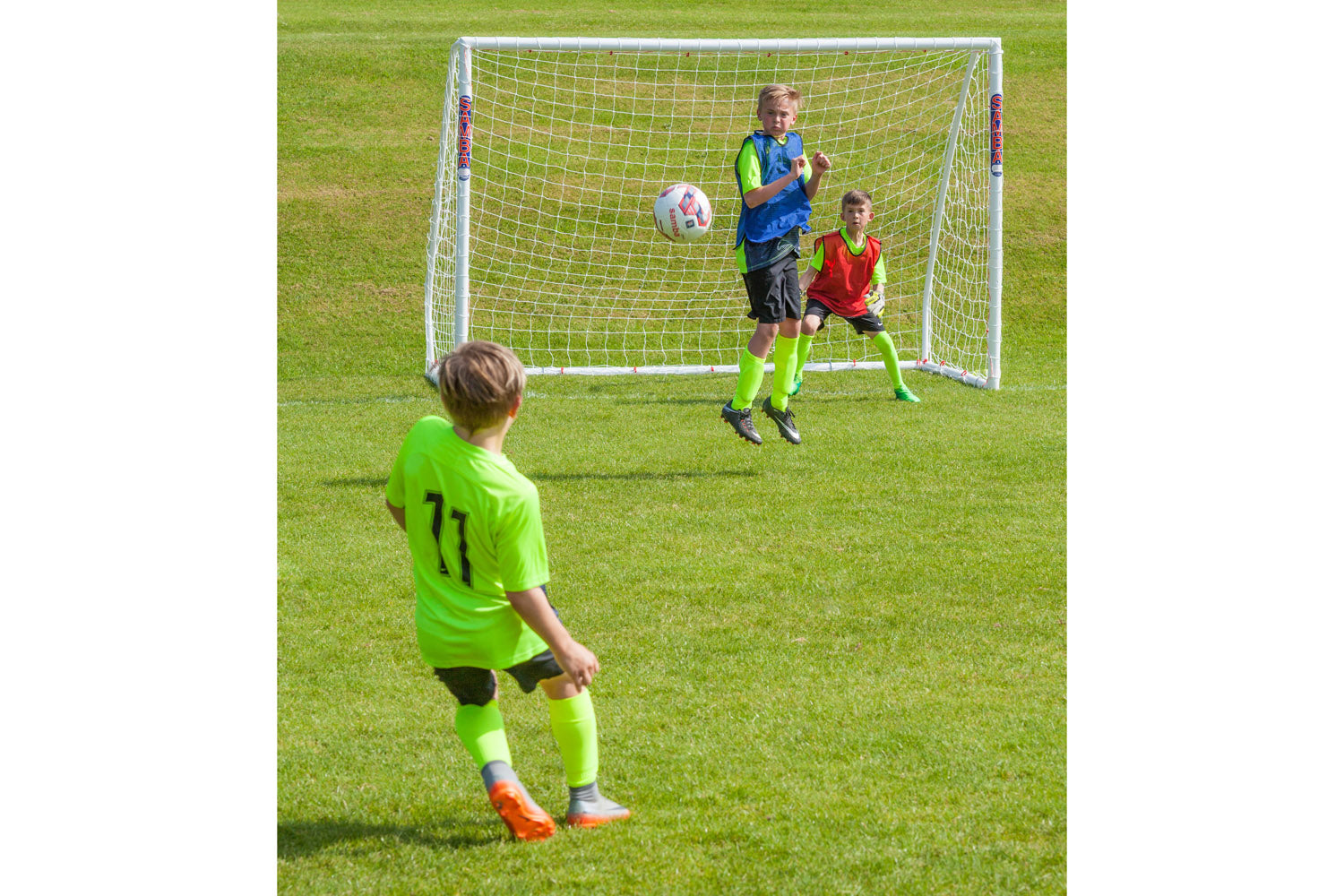A child in a neon green soccer uniform prepares to kick a ball toward a Samba Sports Samba Futsal Match Goal 3m x 2m, as one player jumps to block and another crouches in front of the high impact uPVC goal, both in bright uniforms.