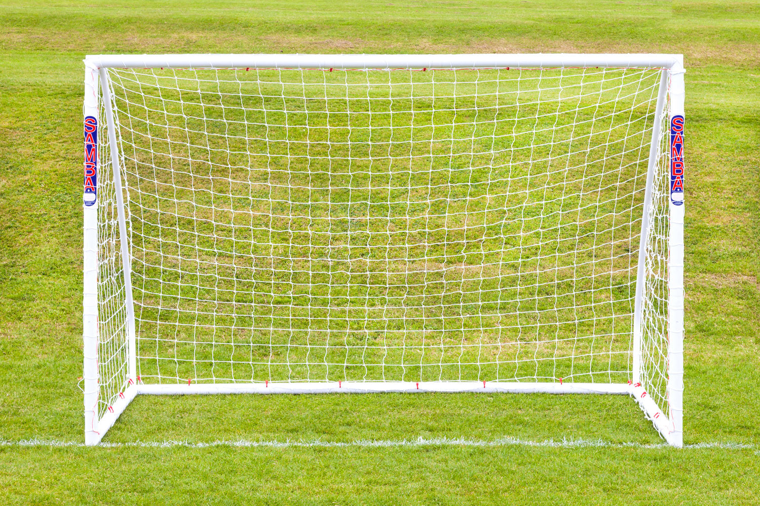 The Samba Sports Samba Futsal Match Goal 3m x 2m, made of white high-impact uPVC and complete with a net, stands on a green grass field marked with white boundary lines in the foreground and background.