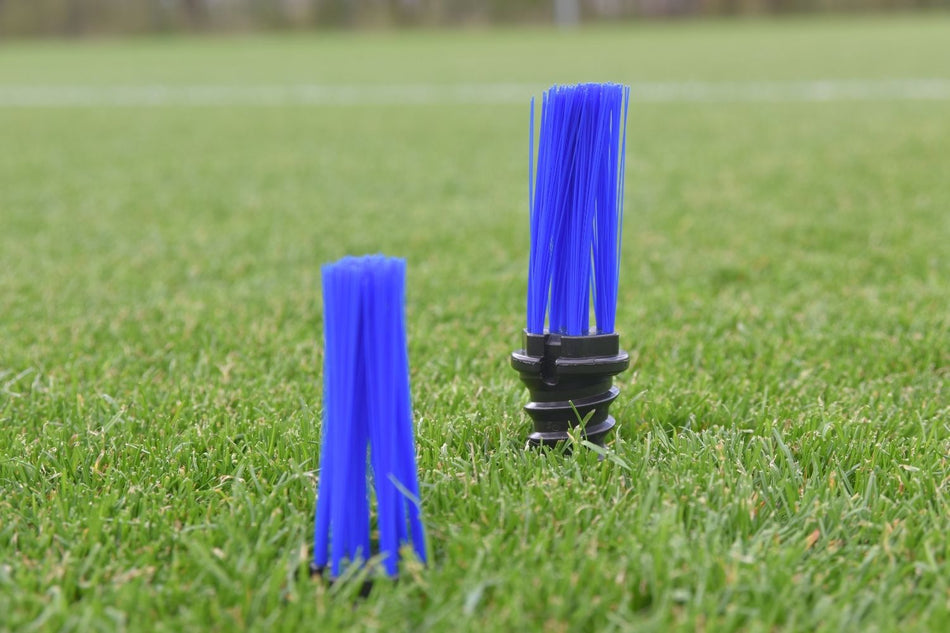 Two blue Samba Grass Tuft Markers by Samba Sports are upright on green artificial turf; the larger marker has a black base, while the smaller sits directly on the turf. A blurred sports field appears in the background.