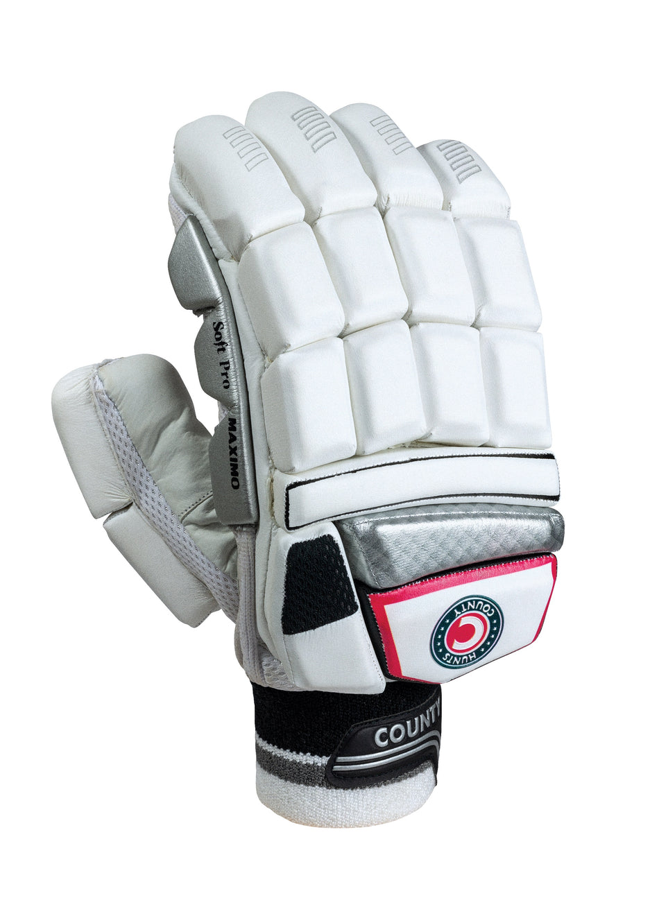 The Hunts County Maximo Batting Gloves by Hunts County feature high density foam split finger protection, a black and red logo patch on the wrist strap, and textured grip on the palm and thumb. The white glove is shown upright against a white background.