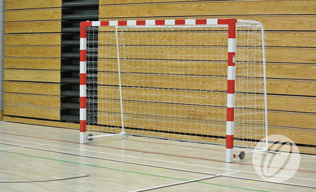 A Harrod Sport indoor soccer goal fitted with HB1 2.5MM Handball Nets White is positioned on a gymnasium floor with wooden panel walls in the background, and features small wheels for easy movement.