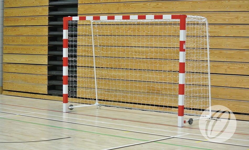A Harrod Sport indoor soccer goal fitted with HB1 2.5MM Handball Nets White is positioned on a gymnasium floor with wooden panel walls in the background, and features small wheels for easy movement.