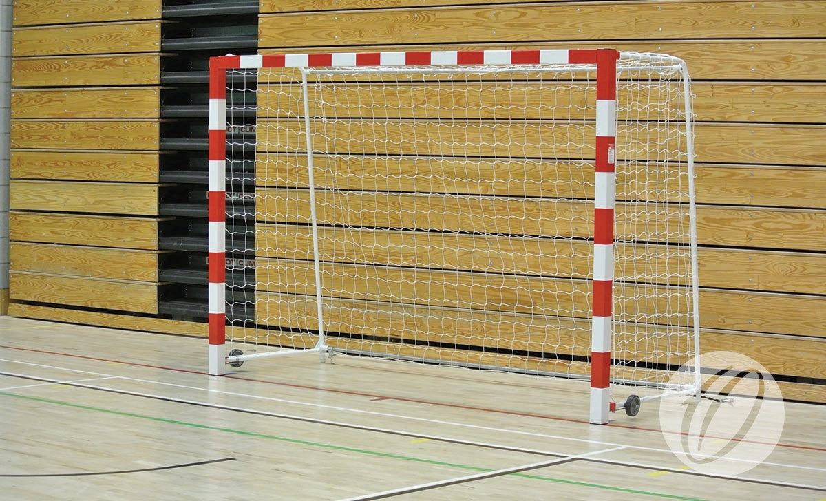 A Harrod Sport Steel Folding Handball Goal with a red and white striped frame, folding side frames, wheels, and attached net is set up on a gym floor against wooden panel walls.