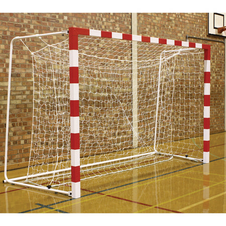 A white and red indoor goal fitted with Harrod Sport's Competition 3MM Handball Nets White stands on a shiny gym floor before a brick wall, with a basketball hoop visible in the background on the right.