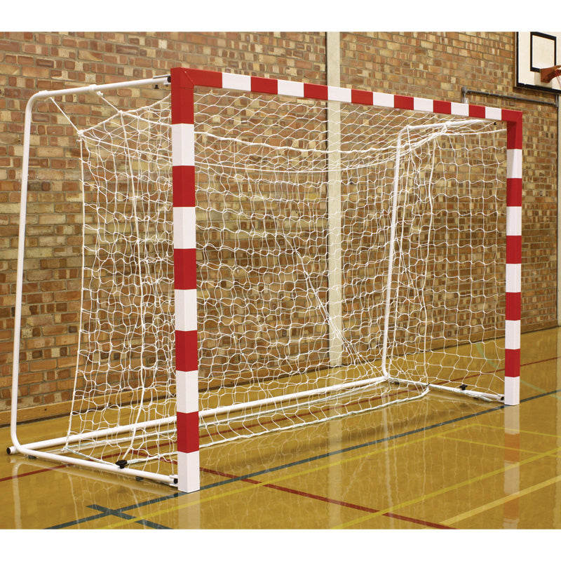 A white and red indoor goal fitted with Harrod Sport's Competition 3MM Handball Nets White stands on a shiny gym floor before a brick wall, with a basketball hoop visible in the background on the right.