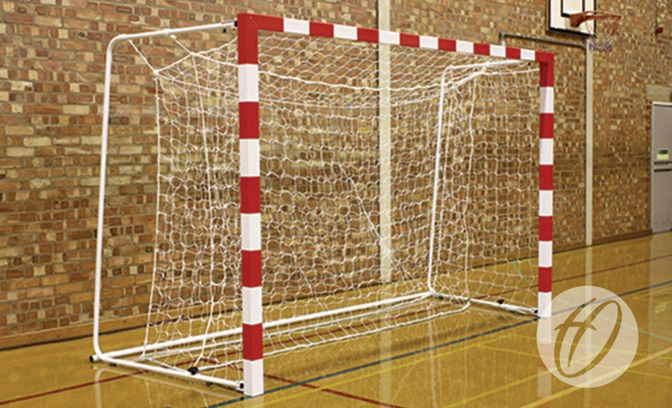 The Harrod Sport Competition Aluminium Handball Goal, featuring red and white stripes and folding net supports, stands in a gym with a wooden floor and brick walls.