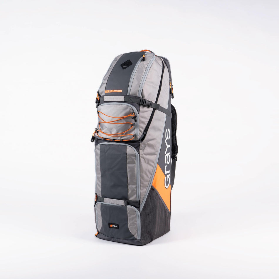 The Grays Alpha-X Kitbag is a large, grey soft-sided hockey bag with orange accents, multiple compartments, a stick pocket, and sturdy handles. "Grays" is printed on the side. The bag stands upright on a white background.
