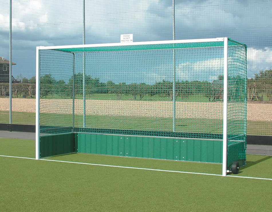 The Harrod Sport Premier Hockey Goals with PVC Backboards, featuring green and white netting, stand on a synthetic surface with a fence, trees, and cloudy sky behind. A small sign is attached at the top of the goal.