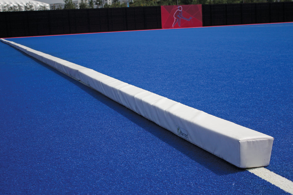 A Harrod Sport Hockey Pitch Divider Pad, long and white with padding, lies diagonally on a bright blue artificial turf field, with a black fence and red banner in the background.