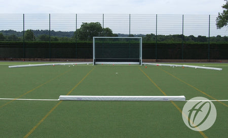A Harrod Sport Hockey Pitch Divider Pad is set up on an outdoor artificial turf field with a field hockey goal, low white barriers, orange cones, a wire fence, and trees and hills in the background.