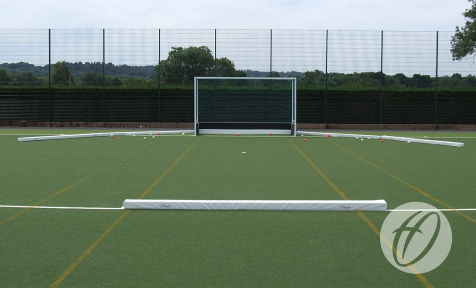 A Harrod Sport Hockey Pitch Divider Pad is set up on an outdoor artificial turf field with a field hockey goal, low white barriers, orange cones, a wire fence, and trees and hills in the background.