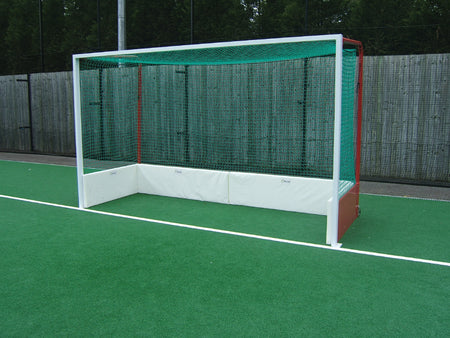 A Harrod Sport Hockey Backboard Protection System with a green net and white frame stands on artificial turf, featuring noise-reducing board protection, with a wooden fence and trees in the background.