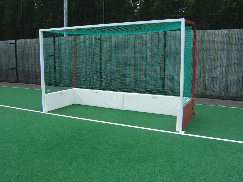A Harrod Sport Hockey Backboard Protection System with a green net and white frame stands on artificial turf, featuring noise-reducing board protection, with a wooden fence and trees in the background.