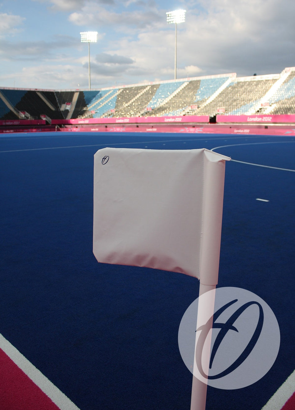 A close-up of the Harrod Sport Single Colour PVC Flag 300x300MM on a blue hockey field in an empty stadium with pink accents and stadium lights, standing under a partly cloudy sky.