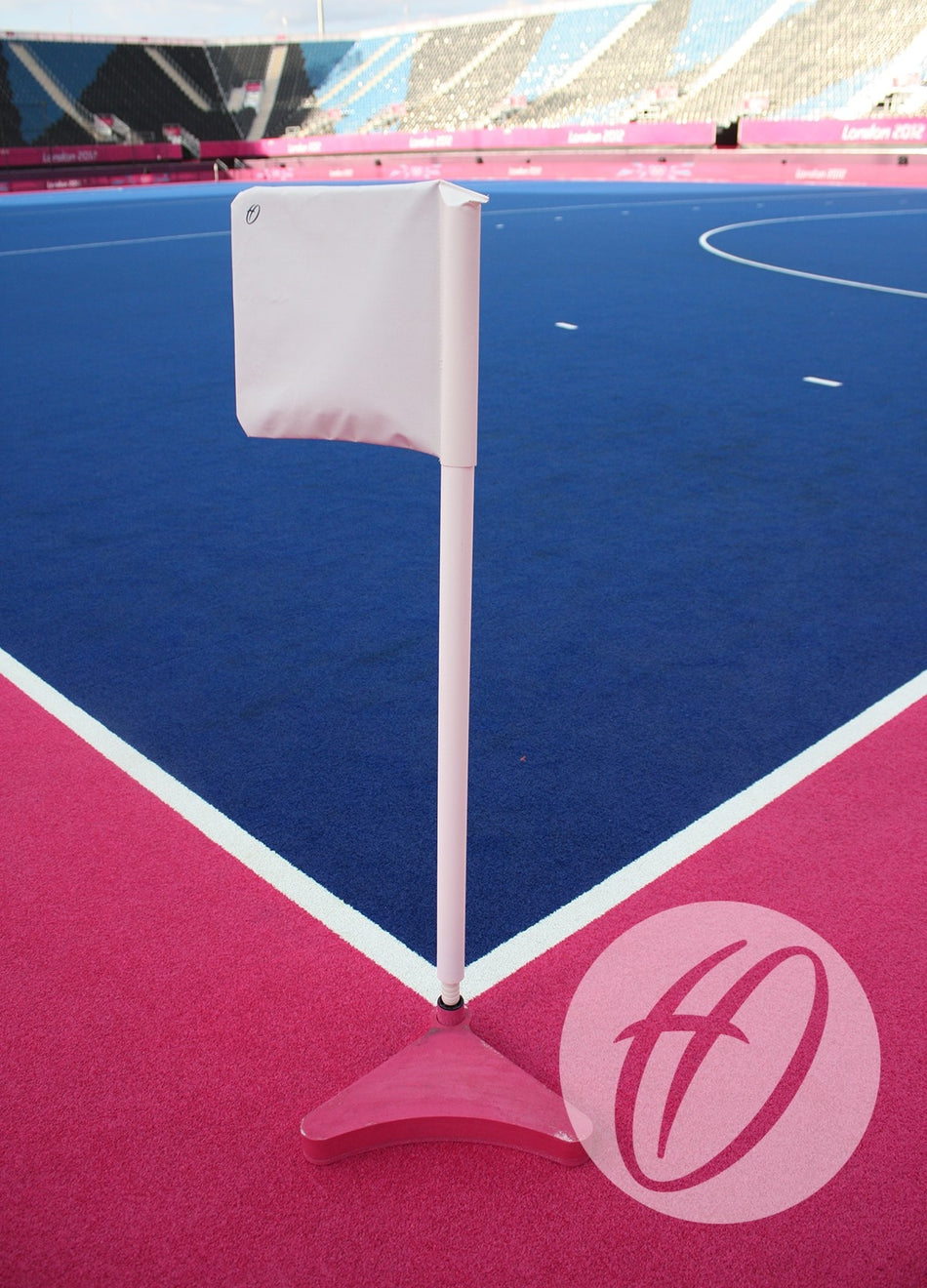 A Harrod Sport Single Colour PVC Flag 300X300MM tops a white field hockey corner post on a pink and blue pitch, with empty stadium seats and part of the London 2012 logo in the background.