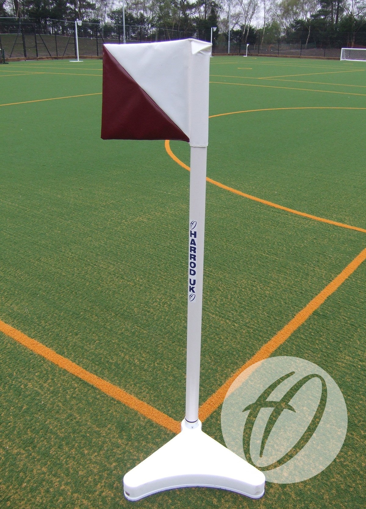 The Harrod Sport 2 Colour PVC Flag 300x300MM (Diagonal) in red and white stands on a white base on outdoor artificial turf with yellow boundary lines, trees, and fencing in the background.
