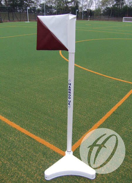 The Harrod Sport 2 Colour PVC Flag 300x300MM (Diagonal) in red and white stands on a white base on outdoor artificial turf with yellow boundary lines, trees, and fencing in the background.