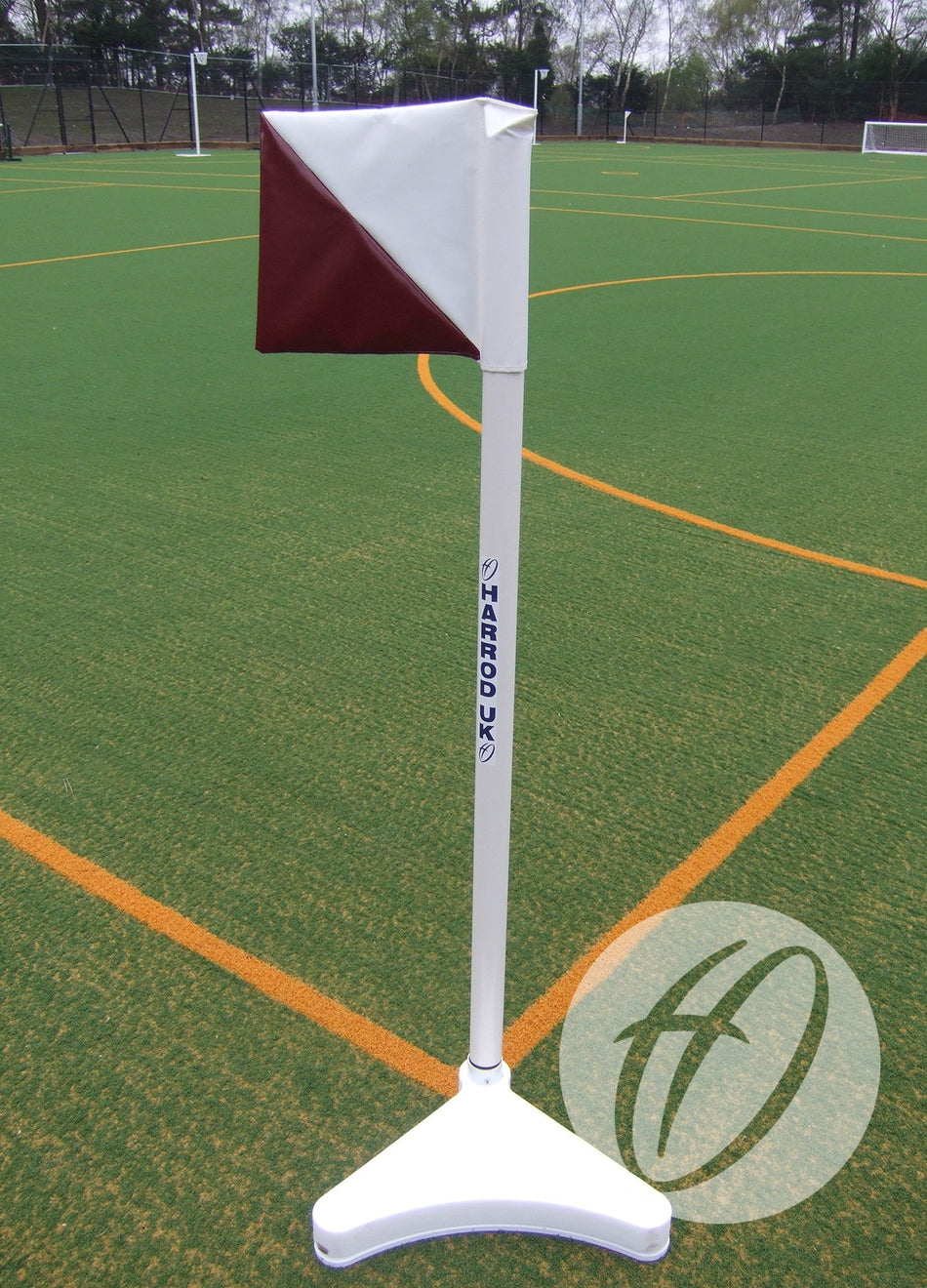The Harrod Sport 2 Colour PVC Flag 300x300MM (Diagonal) in red and white stands on a white base on outdoor artificial turf with yellow boundary lines, trees, and fencing in the background.
