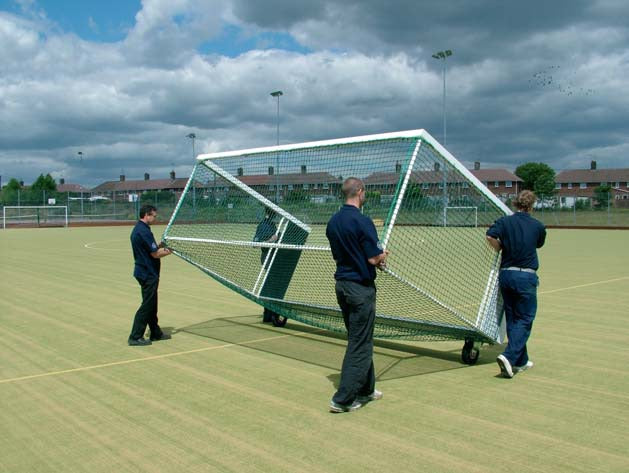 Three people are moving a Harrod Sport Heavy Duty Aluminium Hockey Goal with wheels across an outdoor sports field under a cloudy sky, with houses and trees in the background.