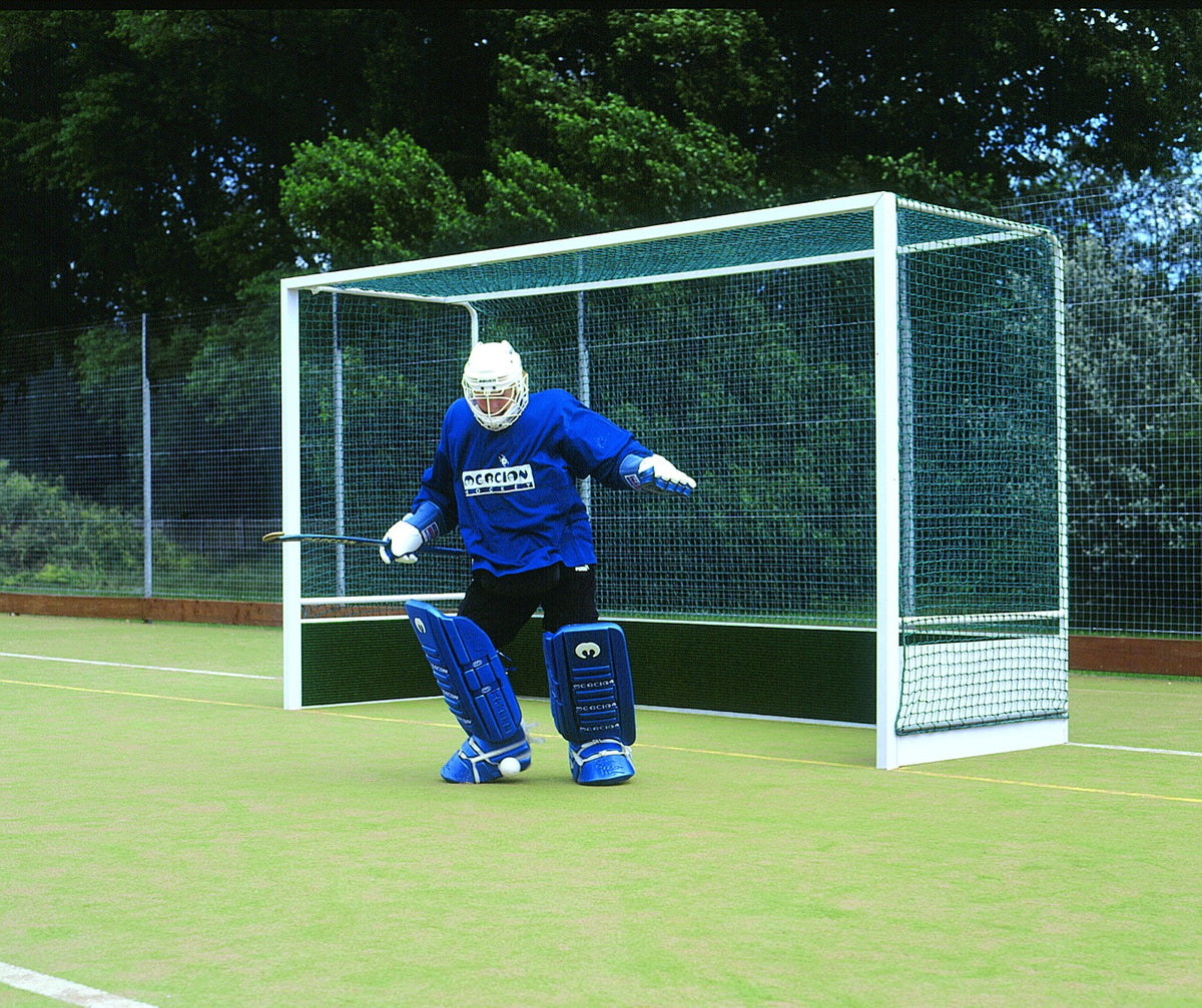 A field hockey goalie in blue gear stands ready to defend in front of a Harrod Sport Heavy Duty Aluminium Hockey Goal on an outdoor field, with trees and a fence visible in the background.