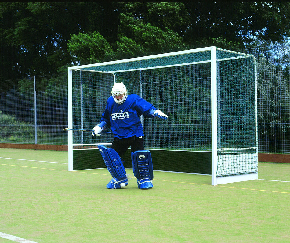 A field hockey goalie in blue gear stands ready to defend in front of a Harrod Sport Heavy Duty Aluminium Hockey Goal on an outdoor field, with trees and a fence visible in the background.