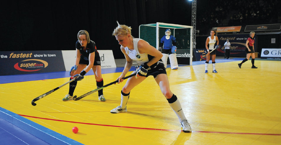 Two female indoor field hockey players compete for the ball near the sideline on a bright yellow court, with a Harrod Sport Heavy Duty Aluminium Indoor Hockey Goal and goalkeeper visible behind them, along with other players near the net.