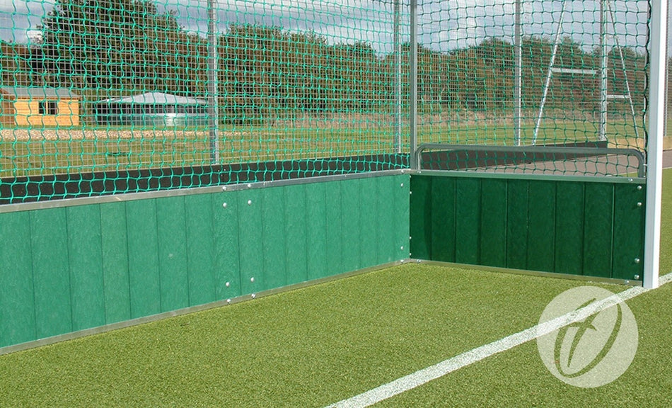 A corner of an outdoor sports field with artificial turf, green fence panels, and netting overhead. White lines mark the field, while Harrod Sport hockey goals fitted with robust PVC Backboards stand ready for play. Trees and buildings can be seen in the background.