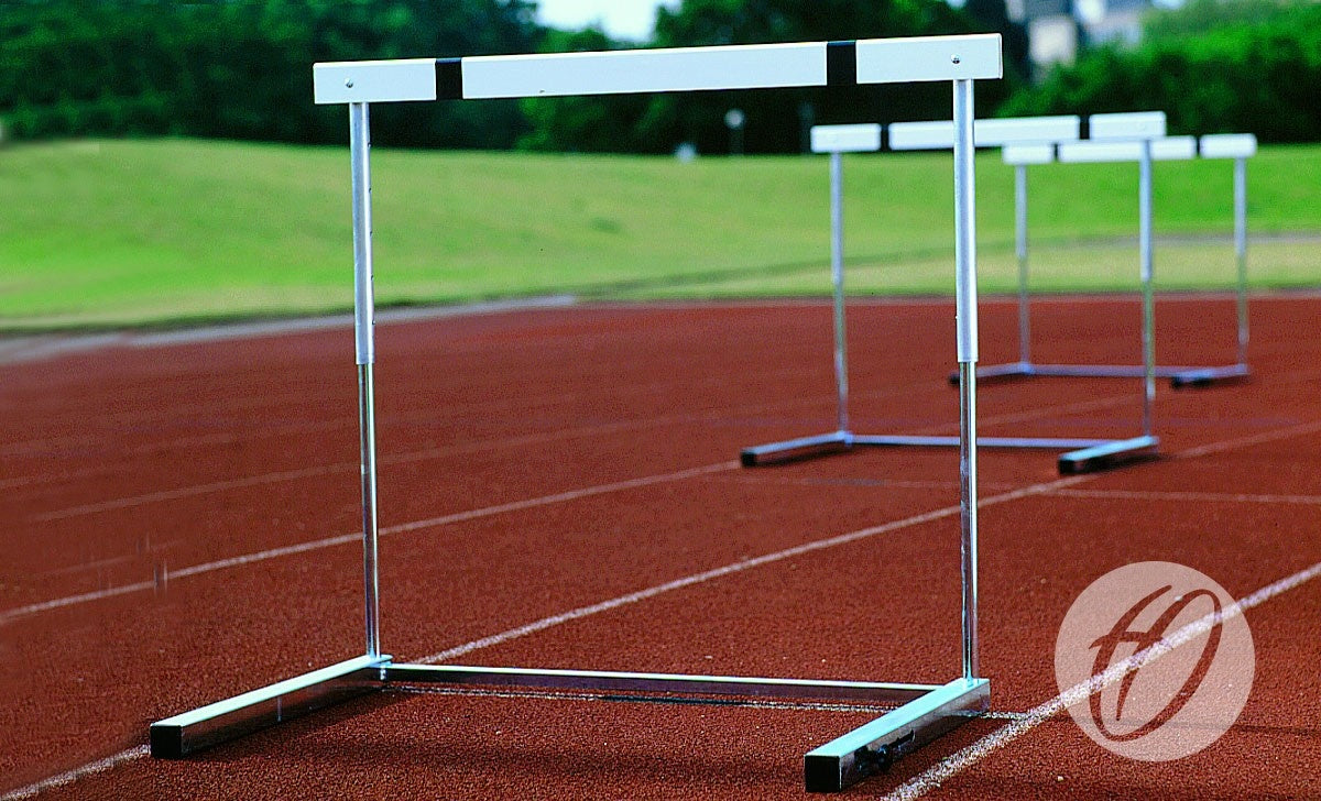 Three Harrod Sport H6 Competition Hurdles, IAAF approved, are set up on a red running track with green grass and trees in the background. The brand logo appears in the bottom right corner.
