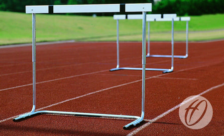 Several Harrod Sport H7 Schools Practice Hurdles are set up on a red running track with grass in the background. The photo highlights the first hurdle, and the Harrod Sport logo is visible in the lower right corner.