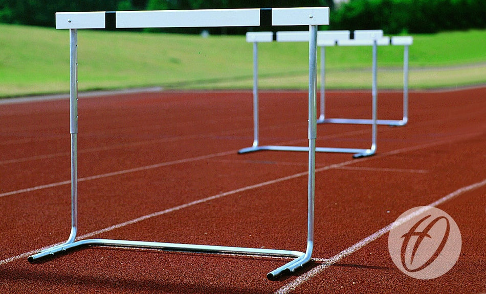 Several Harrod Sport H7 Schools Practice Hurdles are set up on a red running track with grass in the background. The photo highlights the first hurdle, and the Harrod Sport logo is visible in the lower right corner.