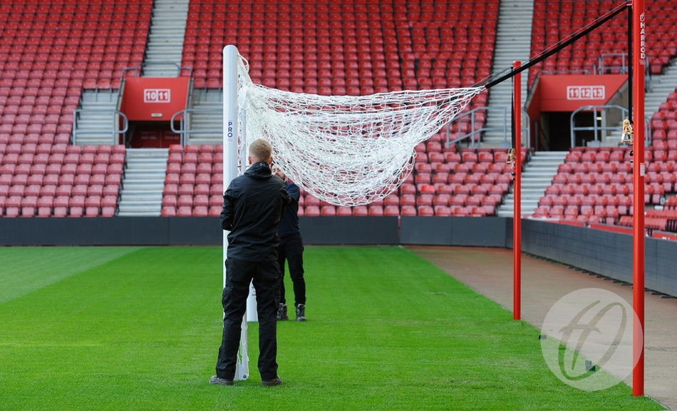 Two people set up a Harrod Sport Stadium Pro Bottom Hinged Net Support with an aluminium net bar on a green field in an empty stadium, rows of red seats visible in the background.