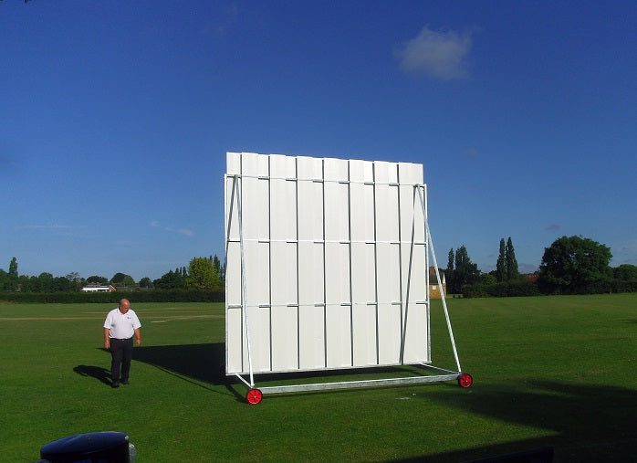 A man stands on grass near a large white Stuart Canvas Polypropylene Sightscreen with red wheels, trees and houses in the background beneath a clear blue sky.