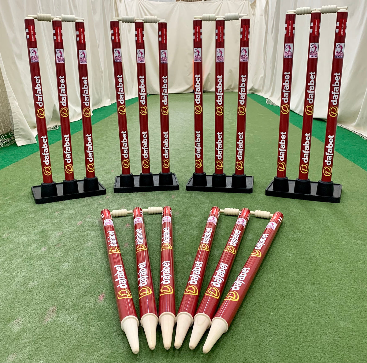 Multiple sets of red Gray Nicolls Custom Printed Match Cricket Stumps with Heavy Rubber Base stand upright in the background, while six more stumps are laid out on the green indoor pitch in front.