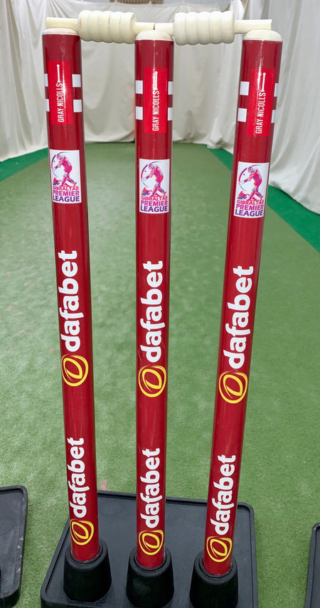 Three red Gray Nicolls Custom Printed Match Cricket Stumps with white handles and heavy rubber bases stand on a green indoor pitch, featuring Womens Premier League 2023 logos. White nets hang in the background.