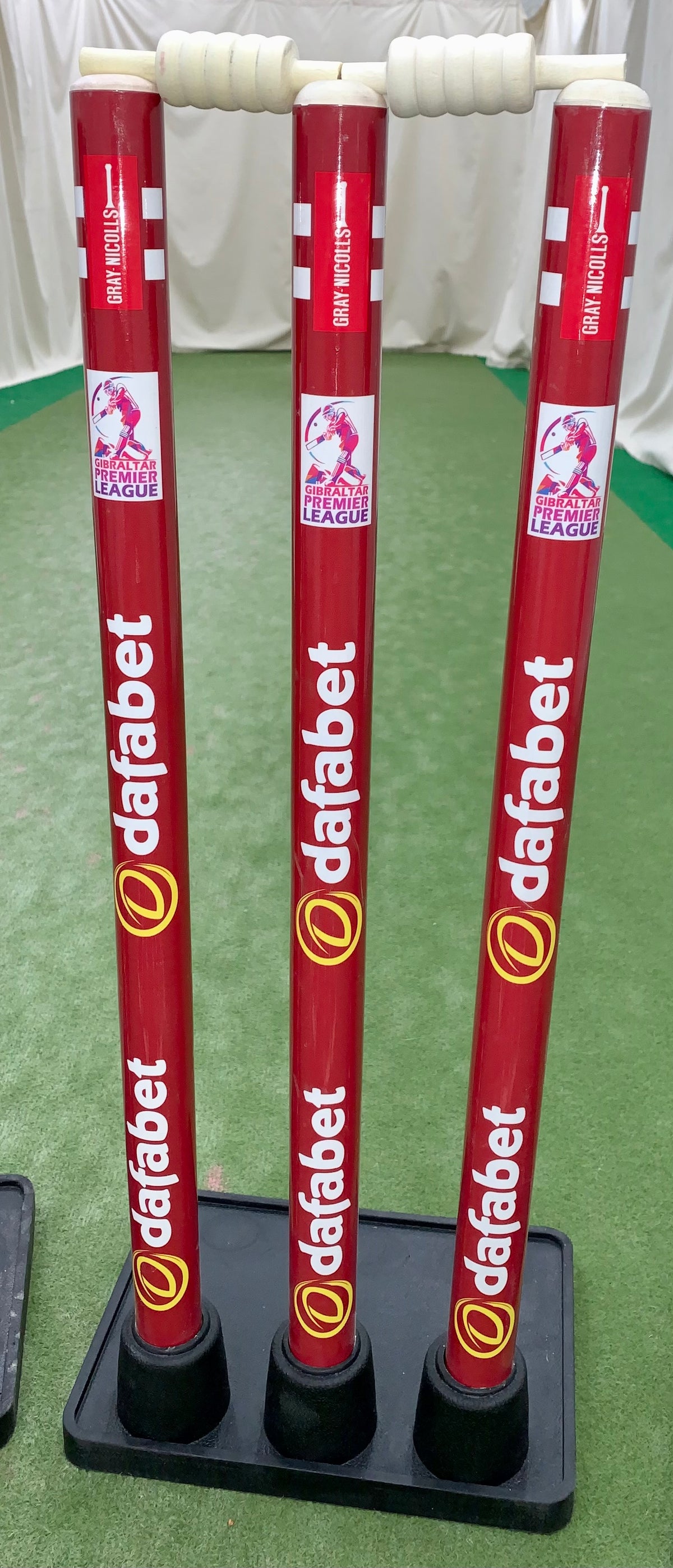 Three Gray Nicolls Custom Printed Match Cricket Stumps with a heavy rubber base stand on a green indoor pitch, featuring white handles and branding, with white curtains in the background.