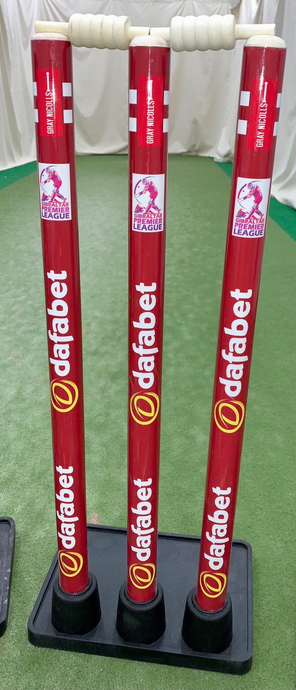 Three Gray Nicolls Custom Printed Match Cricket Stumps with a heavy rubber base stand on a green indoor pitch, featuring white handles and branding, with white curtains in the background.