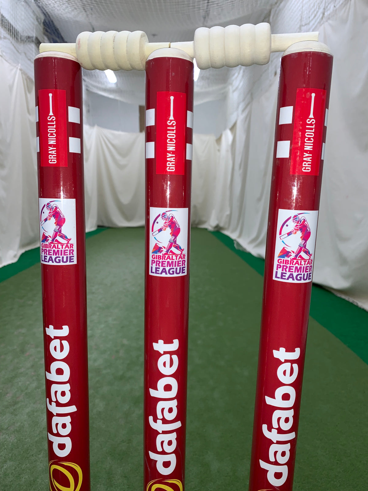 Three red Custom Printed Match Cricket Stumps with Heavy Rubber Base by Gray Nicolls, featuring dafabet and Gibraltar Premier League branding, are set up indoors on green turf with white nets and curtains in the background.