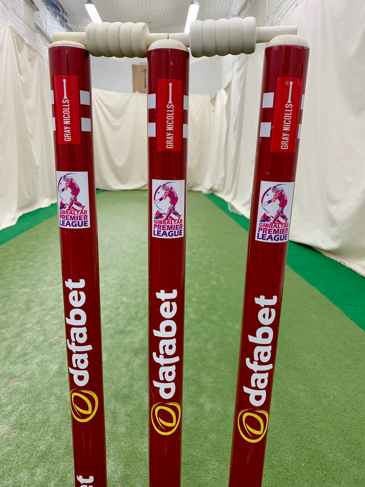Three red Gray Nicolls Custom Printed Match Cricket Stumps with a heavy rubber base and white bails, featuring Dafabet and ShePharo Premier League logos, stand on a green indoor pitch with nets and white walls in the background.