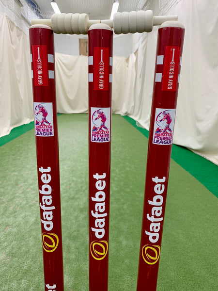 Three red Gray Nicolls Custom Printed Match Cricket Stumps with a heavy rubber base and white bails, featuring Dafabet and ShePharo Premier League logos, stand on a green indoor pitch with nets and white walls in the background.